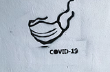 Graffiti on a white wall displaying a mask and “COVID-19” written below it.
