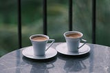 Two cups of coffee sit side by side on a marble table, morning light falling softly across them.