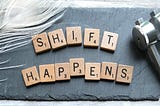 The image shows wooden Scrabble letters on a piece of grey slate, spelling out the words Shift Happens