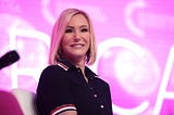 A middle-=aged woman with shoulder-length straight blonde hair, wearing a button-down shirt dress with red, white, and blue ribbons at the collar and sleeves, smiles down from a stage.