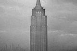 Long shot of the top half of the Empire State Building against a cloudy sky. King Kong falls from the very top, bouncing on the way down.