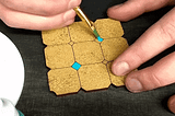 moving gif of white hands painting small cyan diamonds on a sand-coloured grid