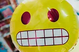 A yellow grimace face emoji balloon in front of a blurry, colorful background.