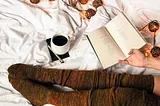 book of poetry with long stockings on a bed with coffee, a pen and lights