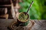 A ceramic cup of mate sits on a wooden table. It is full of a drink with a lot of green herbs floating on the top, and a spoon sticking out of it.