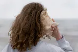 Brown-haired woman with her back to us listens with her right ear to a large seashell.