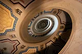 Oval shaped spiral of a staircase from below. Each layer of the staircase has painted patterns in colors of blue, ochre, yellow, brown — all with a centuries old patina. It is clear that it spirals upward several flights.