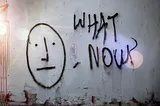 Graffiti with staid face and “What Now?” spray painted on the the wall of an abandoned building.