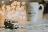 a toy VW campervan on a map on a table, in the background, out of focus, are fairy lights and a mug.