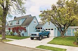 A two-story home painted blue with an attached garage. A blue pickup truck is parked in the drive. The grass and landscaping are very tidy.