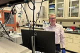 Brad Sutliff wears safety glasses as he stands in the lab in front of a computer monitor.
