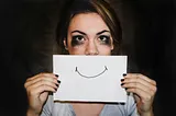 A photo of a woman with smudged eye make up holding a piece of paper with a smile on it in front of her mouth