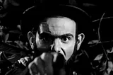 Close up shot in black and white of a man with a full beard, wearing a top hat, and pointing at the camera.