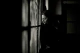 A black and white image of a lone woman in a dark room looking out the window longingly. This demonstrates the isolated feelings associated with agoraphobia and the fear of leaving the house.