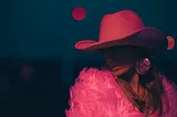 A woman wearing a bright pink cowboy hat and a bring pink feather jacket. The woman has long hair and her face is obscured by the shadow from the hat. The background to the picture is dark except for a bright pink moon.