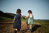 Image shows two young girl children, holding hands, walking away from the camera along a dirt path.