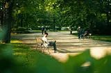 A tree-lined street in a park, a man sitting on a bench with his phone, and a few people strolling.