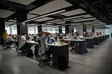 Dozens of people sitting and staring at their laptops on a series of long desks.