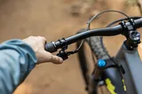 A photo of a man's left hand, reaching down to grasp the left handlebar of a bicycle. The handlebars are black, and we only see the left-hand side of the handlebars, with the front wheel of the bicycle out of focus in the background of the picture.