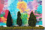 The image shows a vibrant, colorful mural on a wall, with the artwork giving the impression of an abstract sunset with blue sky above and various colors blending into each other. In front of this mural, there are real, three-dimensional shrubs and plants, which include several conical trees and a row of bushy plants with pink flowers, adding a layer of life in contrast to the painted wall. This juxtaposition of living plants against the wall creates a playful interplay between art and nature.