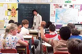 Children sit in a classroom. The teacher stands in front of the chalkboard. The words on the board says, “‘If someone in your family has cancer’ Definition Feelings Treatment.”