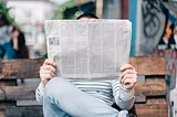 A photo of someone sitting on a wooden park bench, with their legs crossed, holding up a broadsheet newspaper. The newspaper covers their face; only the top of their head, with short black hair, is visible