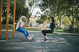 Two smiling women sitting on swings in a city park facing each other.