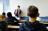 A classroom scene with students seated at desks facing a teacher who is standing at the front of the room near a whiteboard. The students are focused on the teacher, and one student in the foreground has their back to the camera. The classroom has a blue and white color scheme.