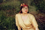 A woman in a bright yellow shirt, glasses and a red head scarf looks off to the side