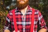 A man with a full red beard and mustache is wearing a red plaid shirt with rolled-up sleeves, red suspenders, and a red bandana tied around his head. He is standing outdoors with a blurred background of trees and foliage. The man has a serious expression and is holding the suspenders with both hands.