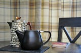 A teapot and a coffee pot sit on a table, in front of an empty chair