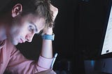 A man at his desk in the dark. He is in front of a computer while looking down at his iPad and holding an Apple Pencil.