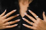 A woman, the image cropped as she displays multiple rings on her hands against a fur coat.
