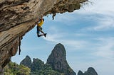 A rock climber holds on (leggs hanging) to a sheer rock face above blue water with verdant islands in the background.