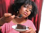 Have some? Person holding a small plate with a slice of black cake — also called Christmas cake or Caribbean fruit cake. The other hand holds a fork with a bite of the cake to share.