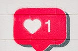 A hand holding a smartphone under a painting of a social media heart “love” symbol.