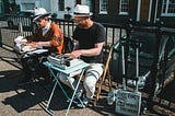 Two men writing on typewriters in the park. There is a fence and a road behind them. One man is wearing white pants, a black shirt, and a hat. The second man is wearing black pants, an orange shirt, a scarf, and a hat.