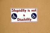 Sign posted on a bulletin board. It sports two drawings of stick figures in wheelchairs along with the words, “stupidity is not a disability.”