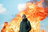 Man in green jacket dark pants, hands in pocket, wearing a grey beanie, standing in front of explosion fire.