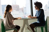 Two women in casual clothing sitting across from each other at a table engaged in conversation