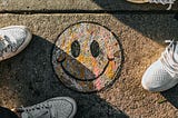 A smiley face that has been chalked onto the sidewalk, with the sneakers of people around it. Photo taken from above.
