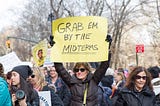 In a group of protestors a woman holds up a sign saying, “Grab em by the midterms.”