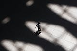 In a black and white photo, a person is seen from a bird’s eye view walking through a scene in which large shadows are cast from an unknown source.