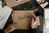 Someone is writing “Welcome” in cursive with a black pen on brown paper. Glasses, a mug, and other papers are sitting nearby.