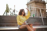 A woman of color is sitting on outdoor steps, smiling and waving. Her laptop and a cup of coffee are in her lap.