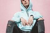 A man sits on the floor with his back against a pink wall, looking disinterested and disinclined to move