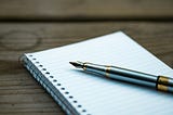 An empty notebook sits on a wooden desk, with a pen sitting atop it.