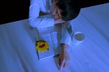 A man sleeping on a desk with an open book, a sunflower, and a white cup with some spilled water in front of him
