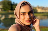 Anika poses in front of a lake on a sunny day, wearing a beige hijab and matching top.