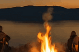 A gif of a fire on the shores of a lake, people sit around looking towards the starry sky, the words “be love” are above the mountain